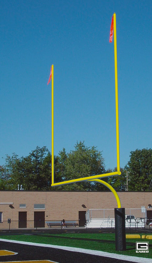 Yellow football goalpost on a field with a building and trees in the background under a blue sky.
