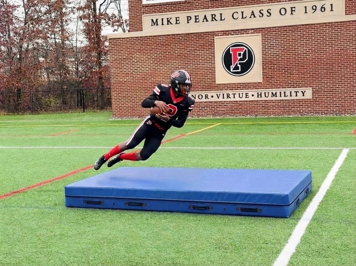 Blue football crash pad on turf as a player in a black and red uniform dives toward it
