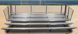 Five-row aluminum bleacher with chain-link fence railing on a dirt field with ocean in background.