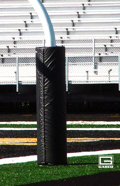 Black football goalpost pad on a field with stadium bleachers in the background.