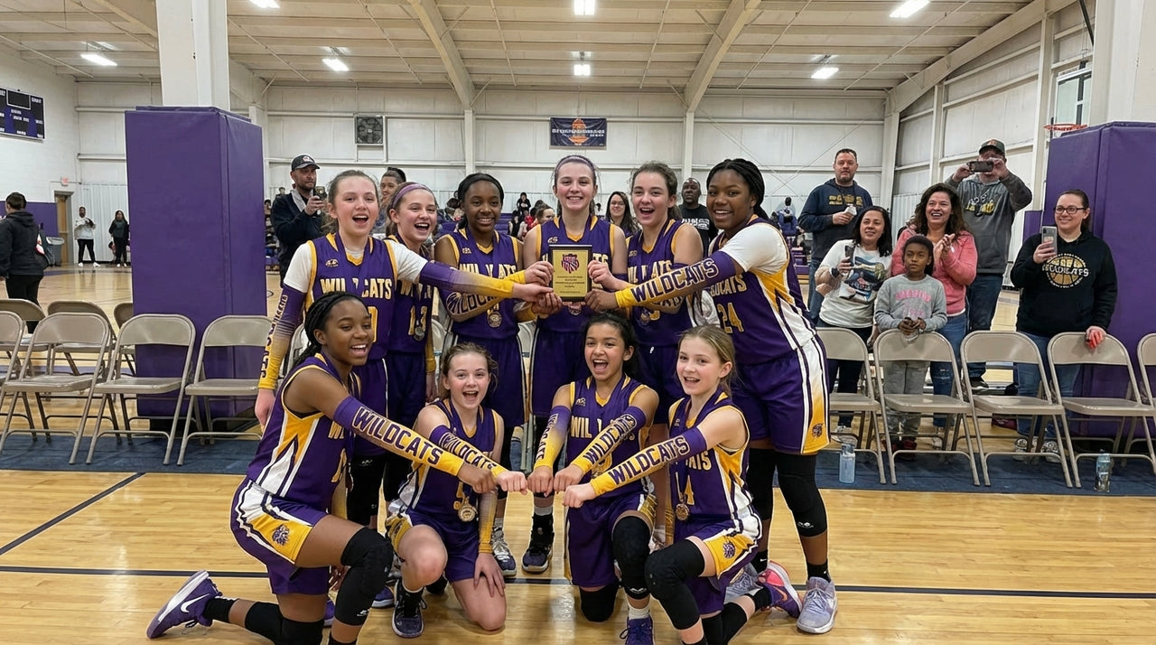 A girls' basketball team in purple and gold uniforms celebrates with a trophy on a court.