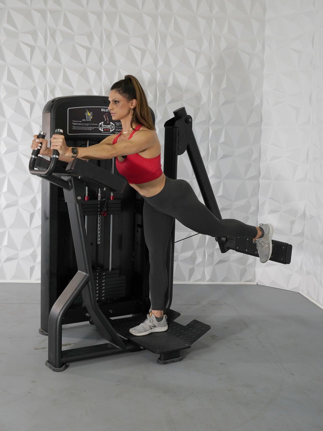 Woman in red top and black leggings using a black glute exercise machine, extending her right leg back.
