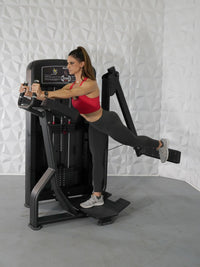 Thumbnail for Woman in red top and black leggings using a black glute exercise machine, extending her right leg back.