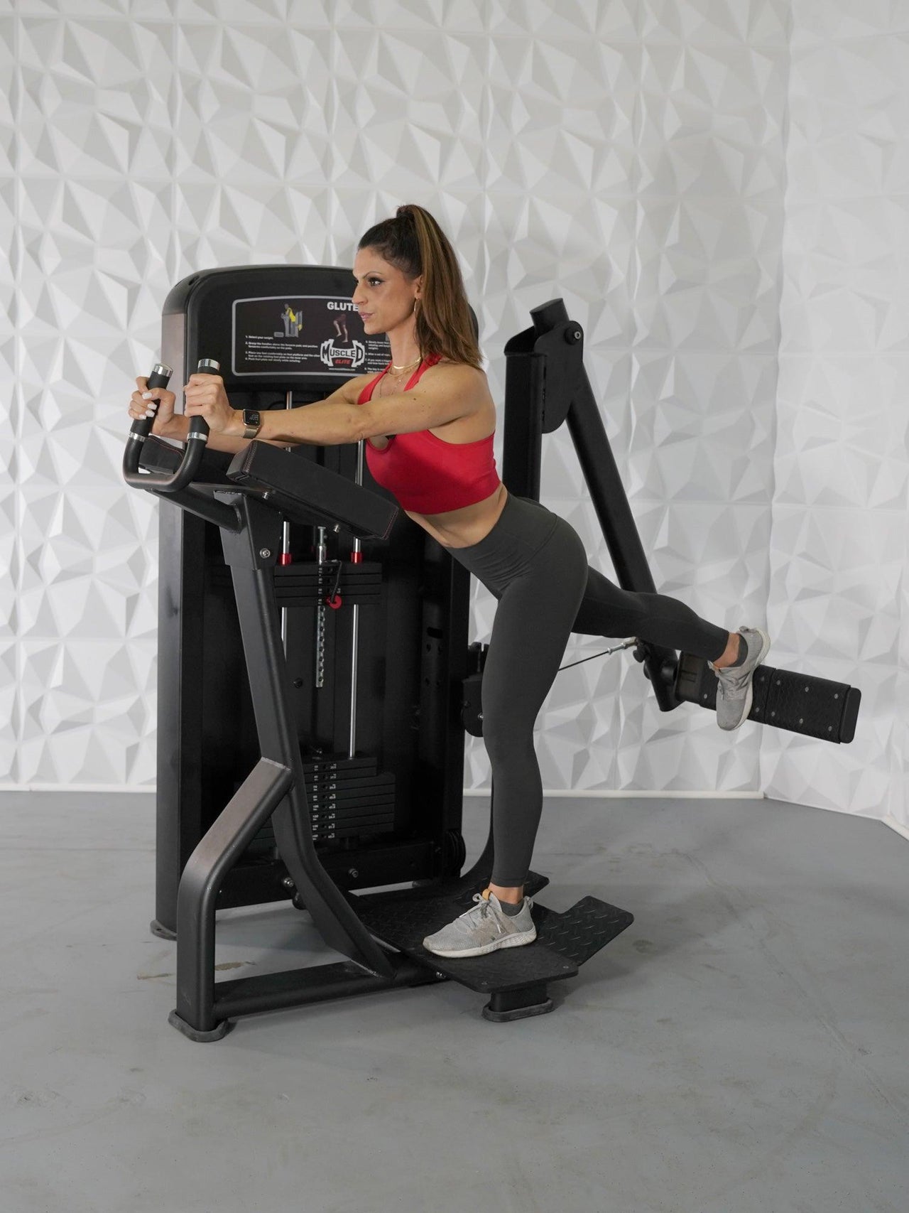 Woman in red top and gray leggings using a black glute exercise machine.