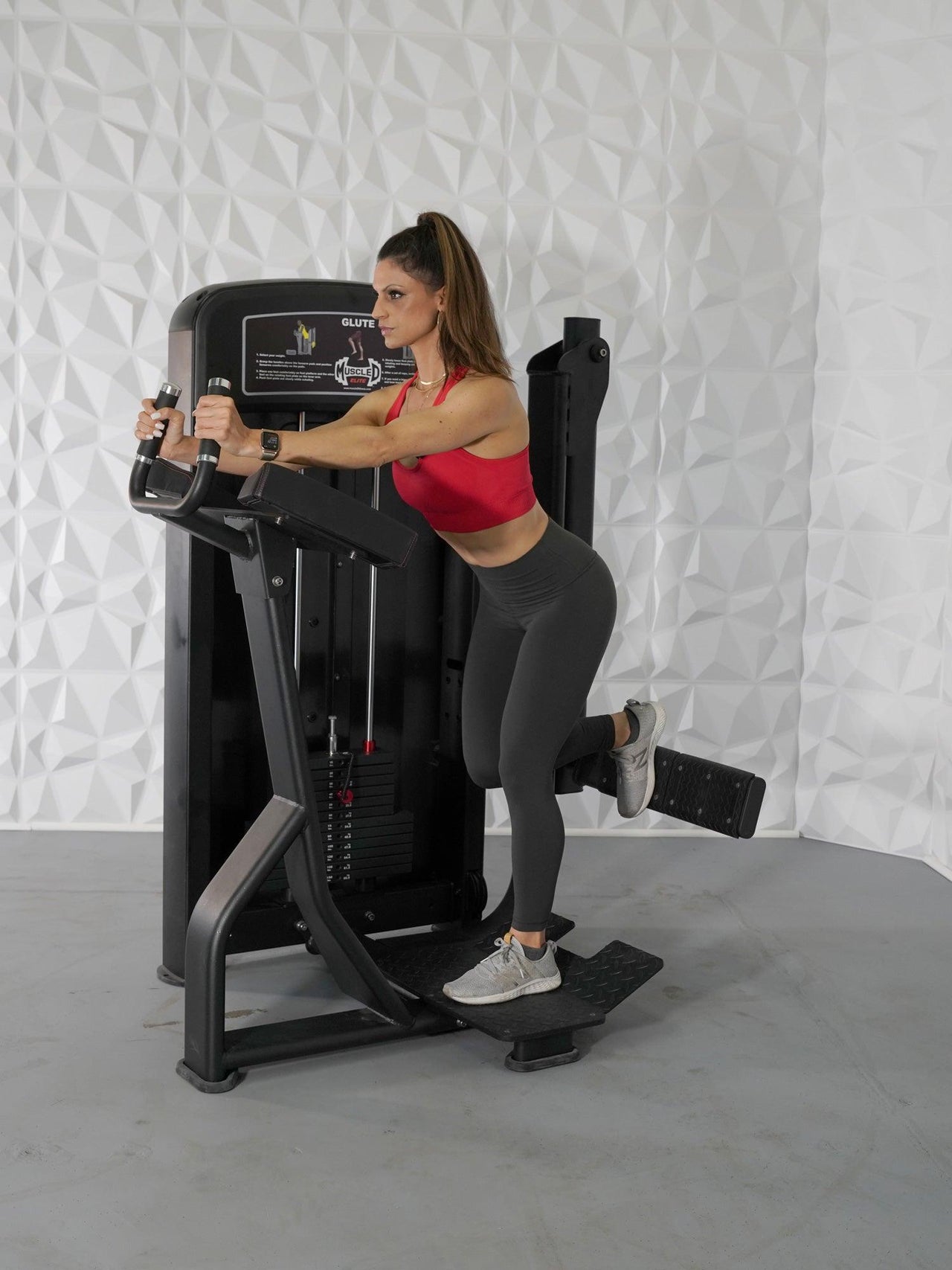 Woman in red top and gray leggings using a black glute exercise machine.