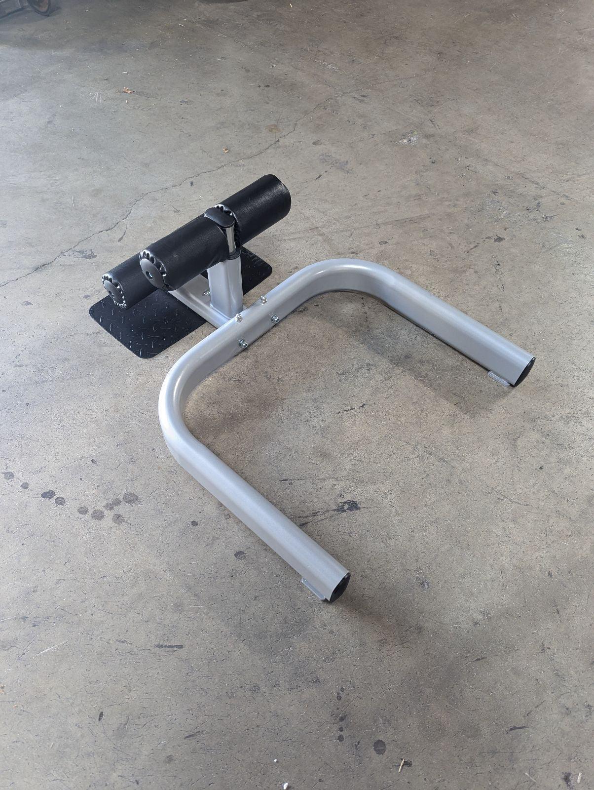 Sissy squat machine with a gray frame, black footplate, and black padded leg rests on a concrete floor.