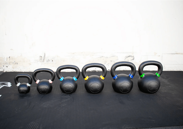 Six black kettlebells of varying sizes with colored bands on handles, arranged on a black mat.