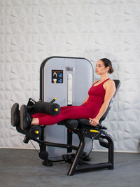Thumbnail for Woman in red using a leg extension/curl machine with black padding and yellow accents.