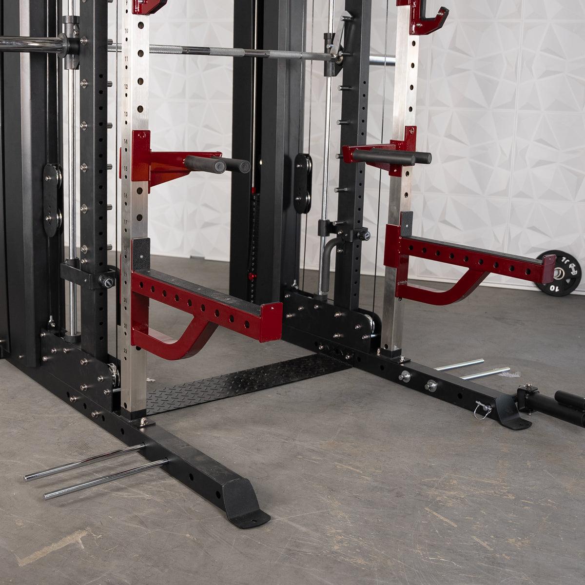 Close-up of a black and red multi-gym machine with a barbell, safety arms, and weight plate storage.