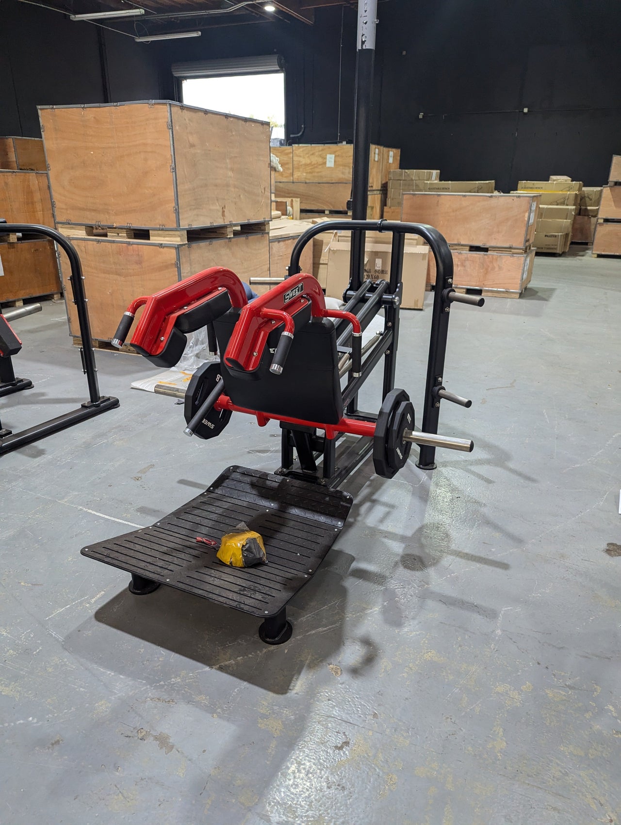 Black and silver hack squat machine with red shoulder pads and weight plates in a warehouse.