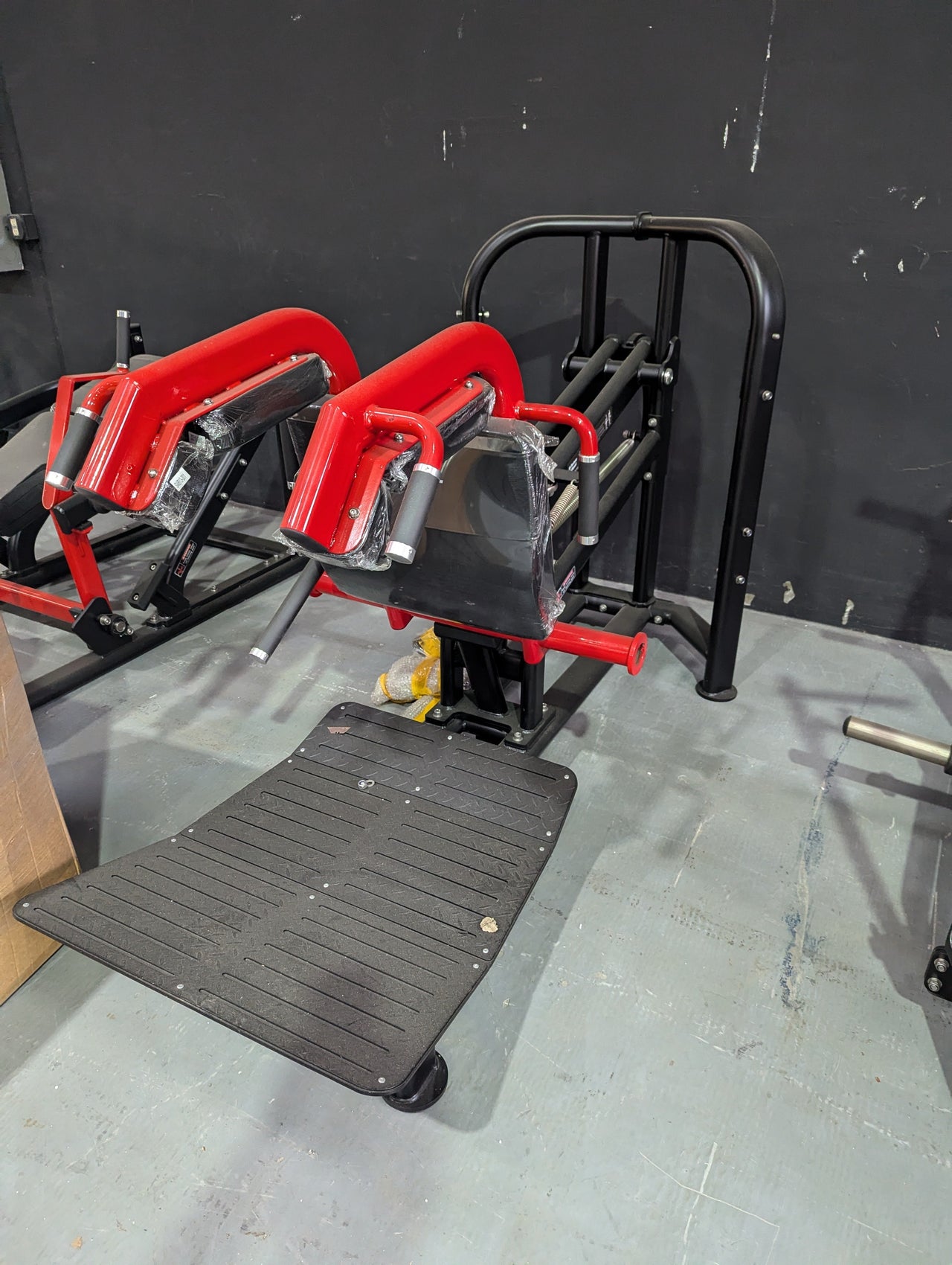 Black and red hack squat machine with a textured footplate against a dark wall.