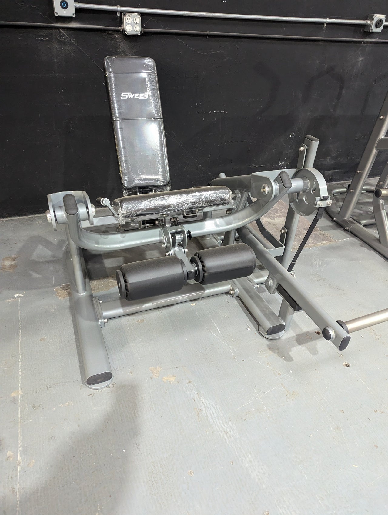 Silver and black Sweet Leg Extension machine with padded seat and rollers, wrapped in plastic.