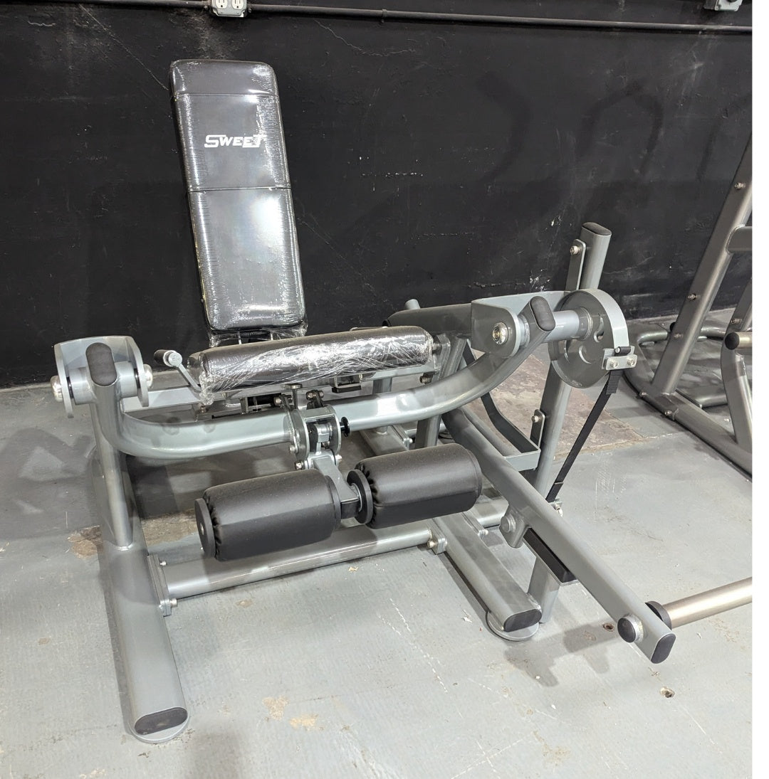 Gray leg extension machine with black padded seat and rollers, some wrapped in plastic, on a concrete floor.