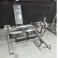 Thumbnail for Gray leg extension machine with black padded seat and rollers, some wrapped in plastic, on a concrete floor.