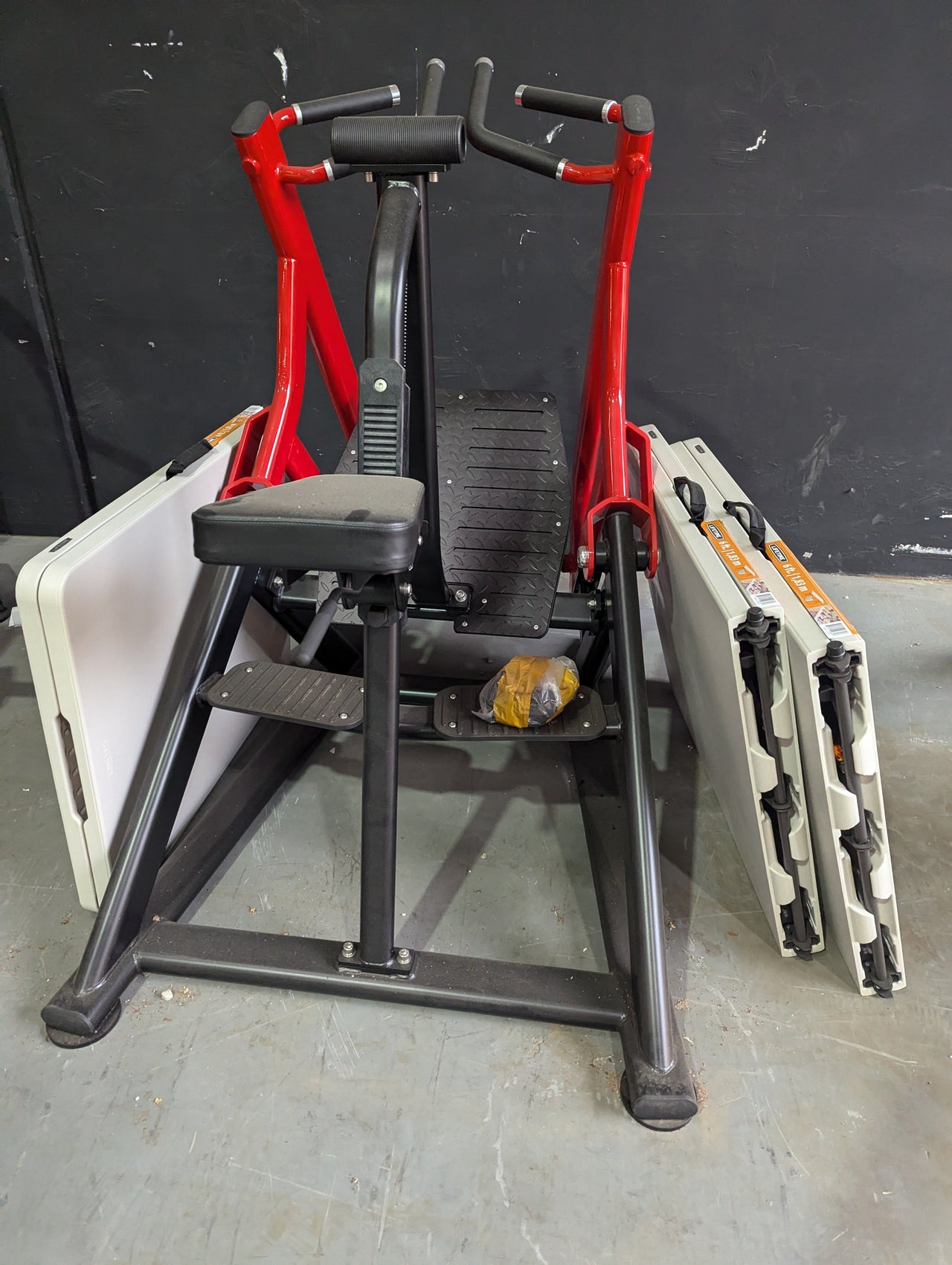 Red and black seated row machine with a padded seat, footrests, and two folding tables leaning against it.