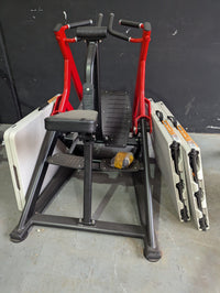 Thumbnail for Red and black seated row machine with a padded seat, footrests, and two folding tables leaning against it.