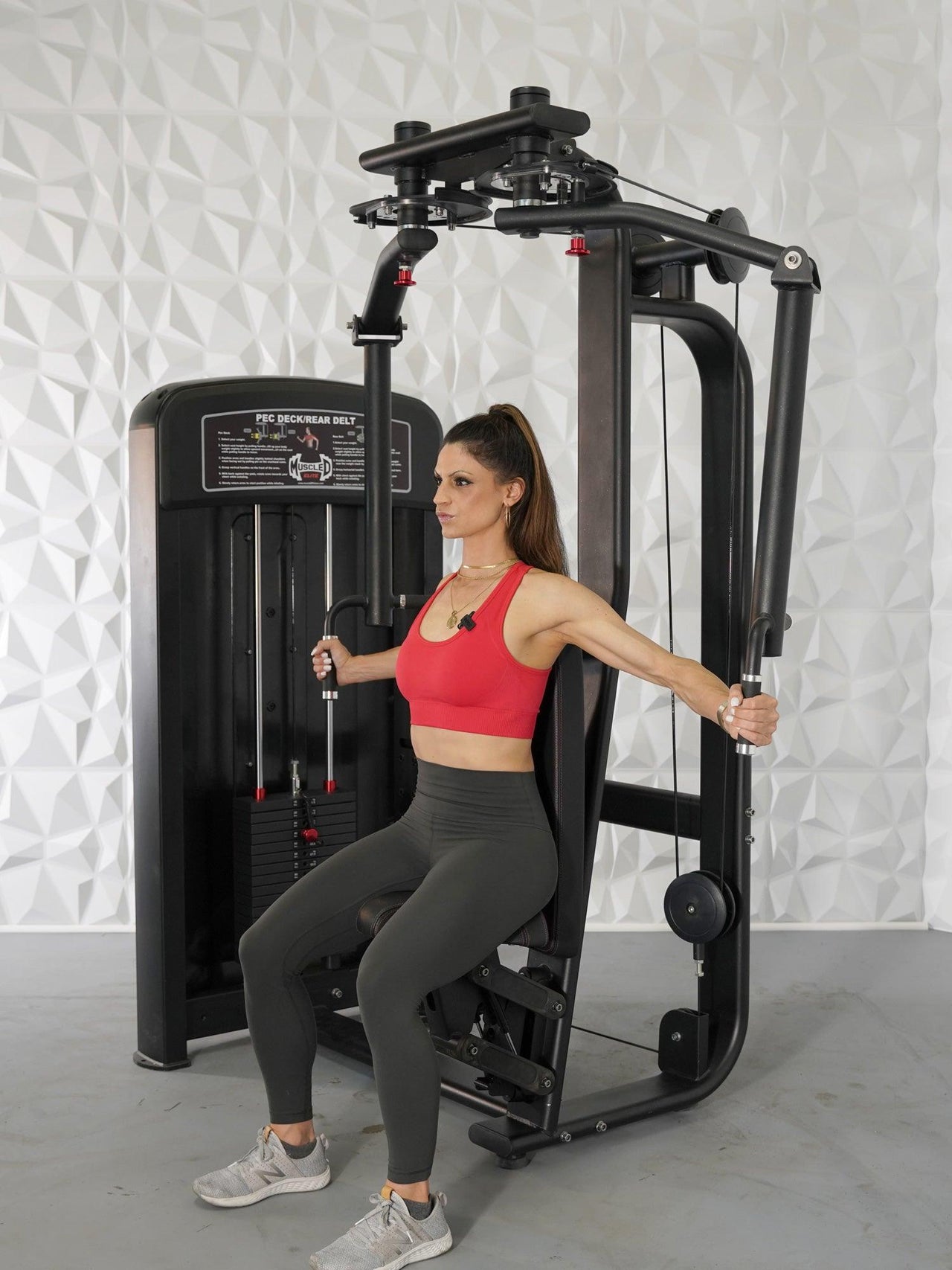 Woman in red top and gray leggings using a black pec deck/rear delt machine.