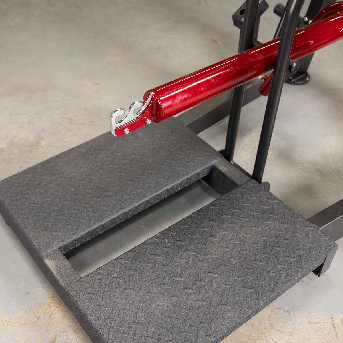 Close-up of the black diamond plate foot platform and red lever arm with hooks on a belt squat machine.