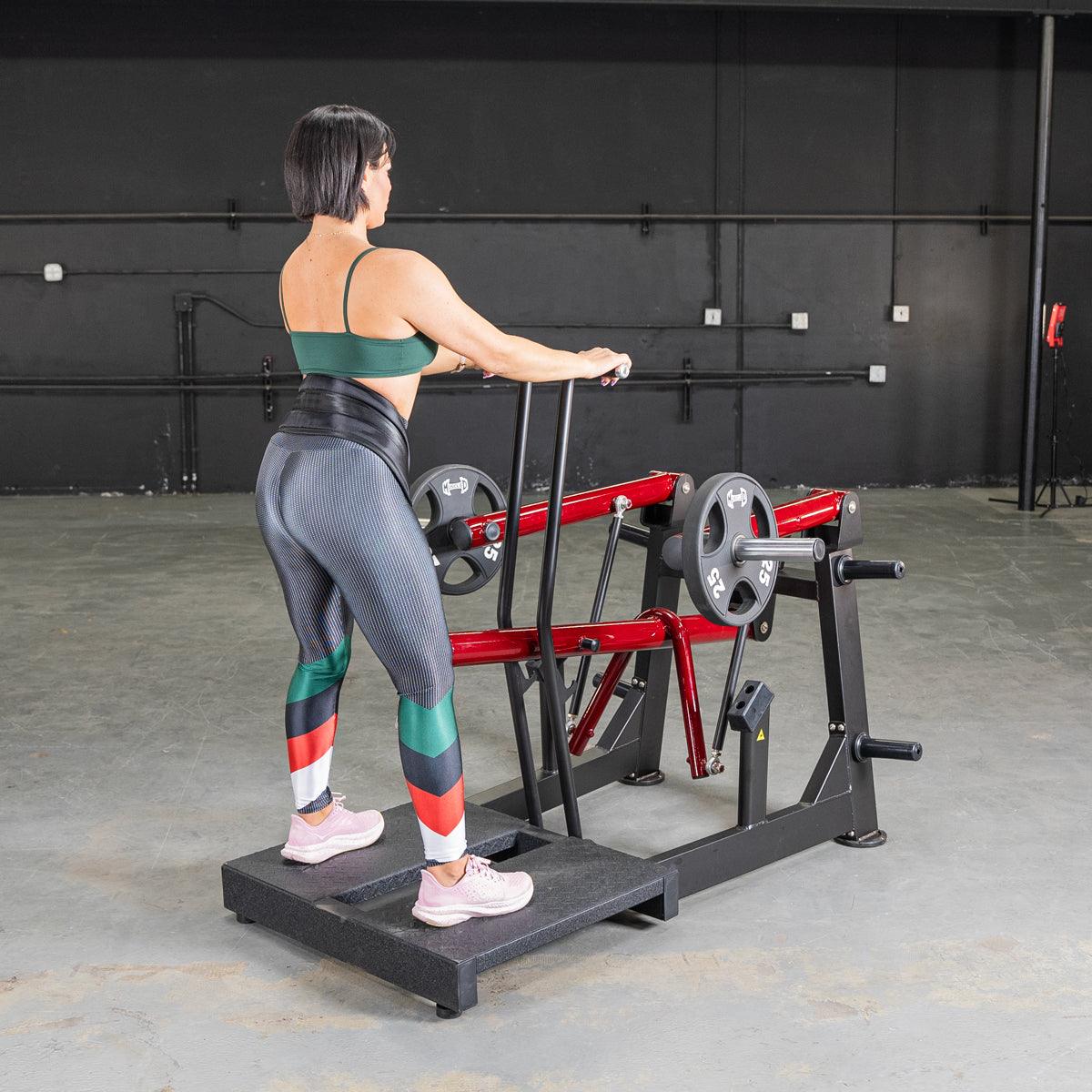 Woman using a black and red belt squat machine with weight plates in a gym.