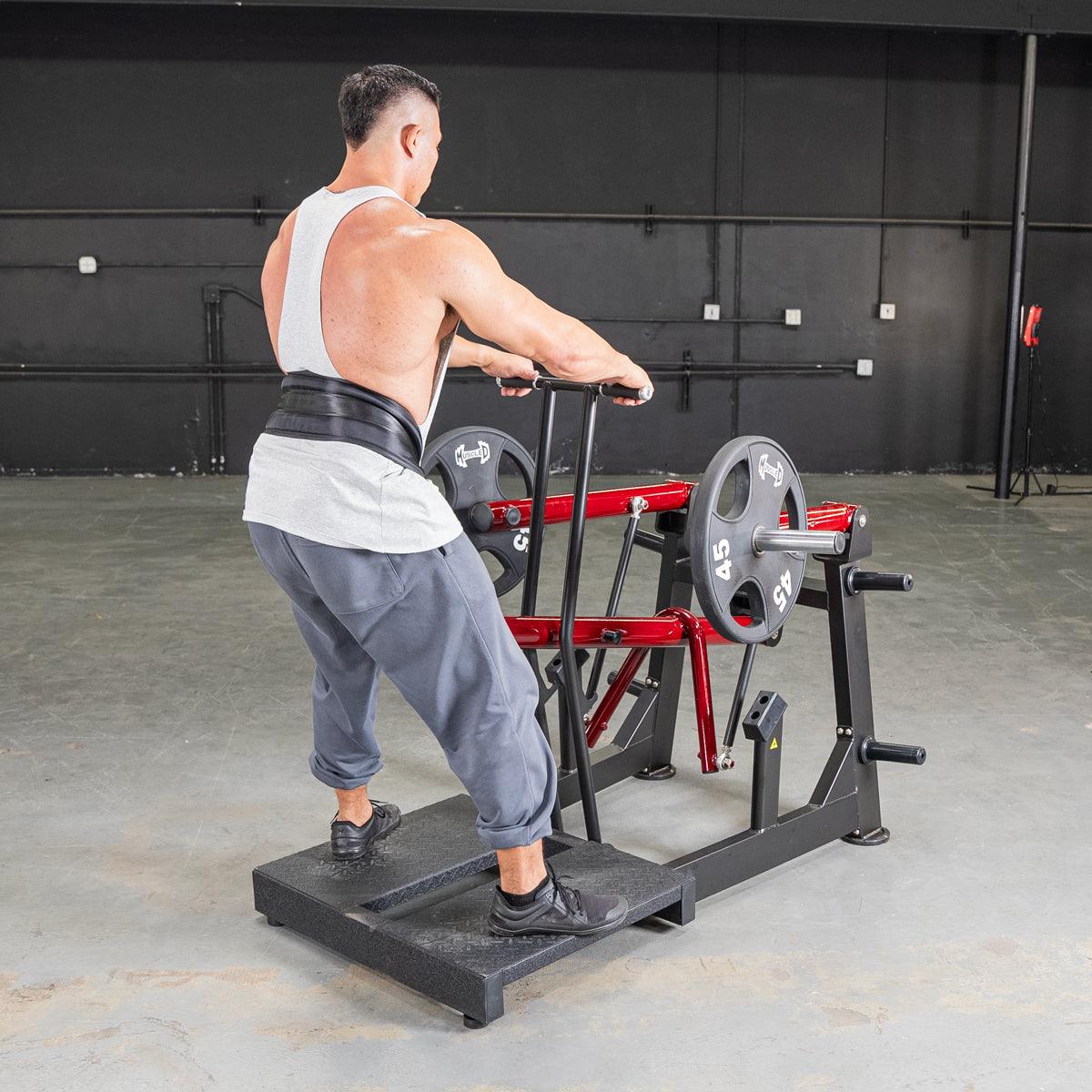 Man in white tank top and weight belt using a red and black belt squat machine with weight plates.