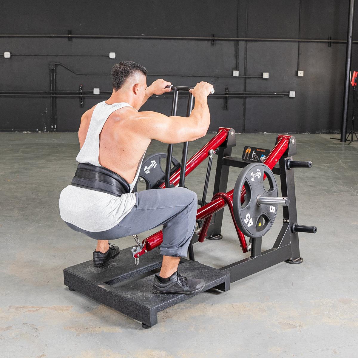 Man in a gym using a red and black belt squat machine with weight plates.