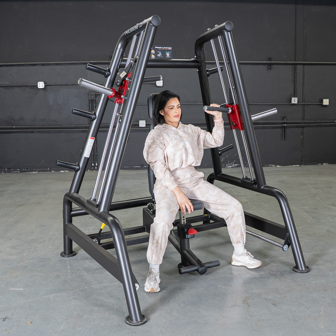 Woman seated on a black leverage bench machine, holding handles for a shoulder press exercise.