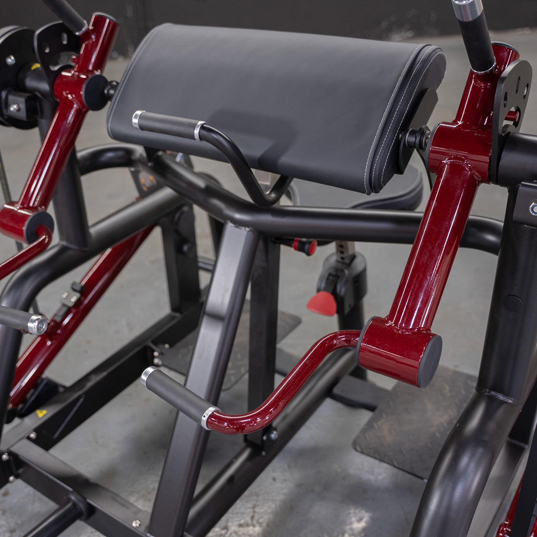 Red and black biceps curl machine with padded armrest and handles.