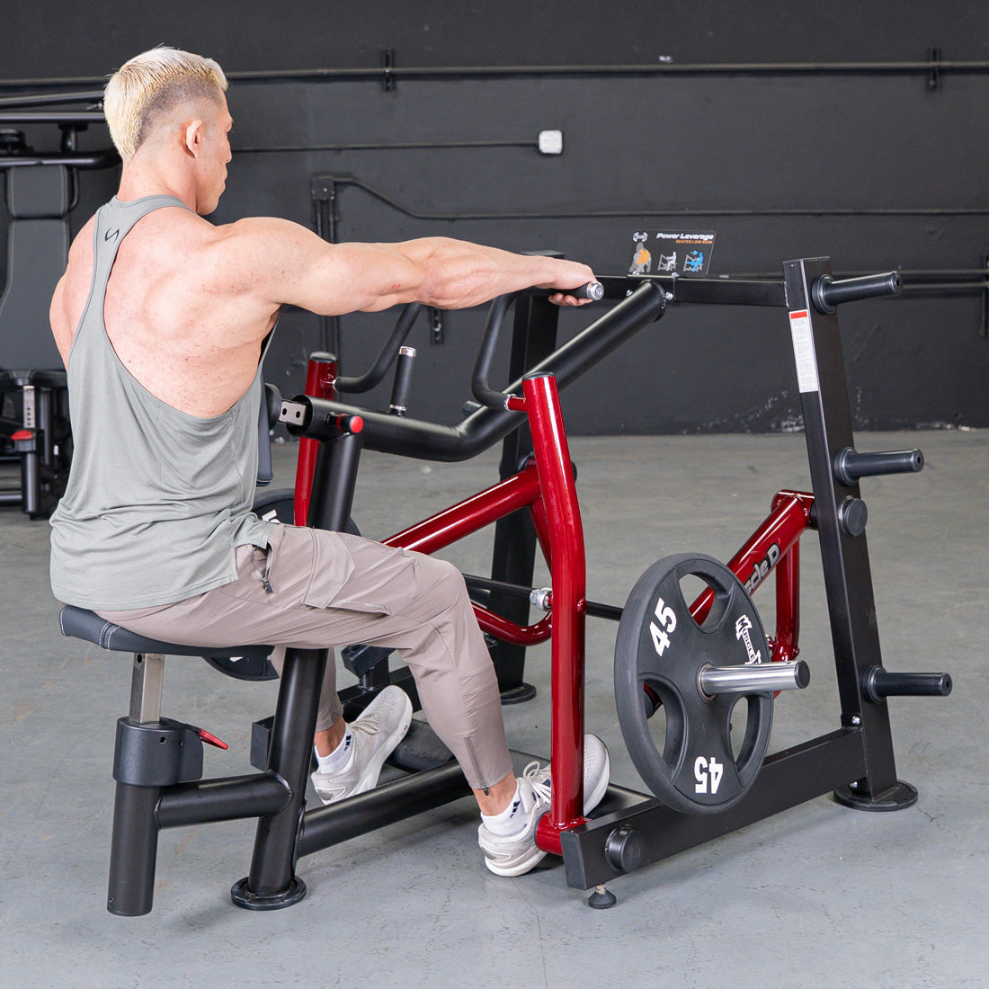 Man in a grey tank top using a red and black seated row machine with a 45lb weight plate.