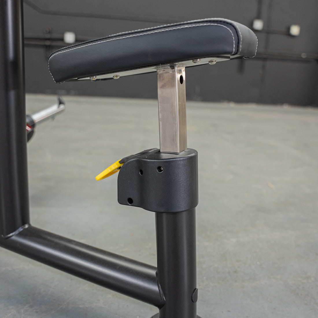 Close-up of a black padded seat with white stitching, adjustable height mechanism, and yellow lever on a fitness machine.