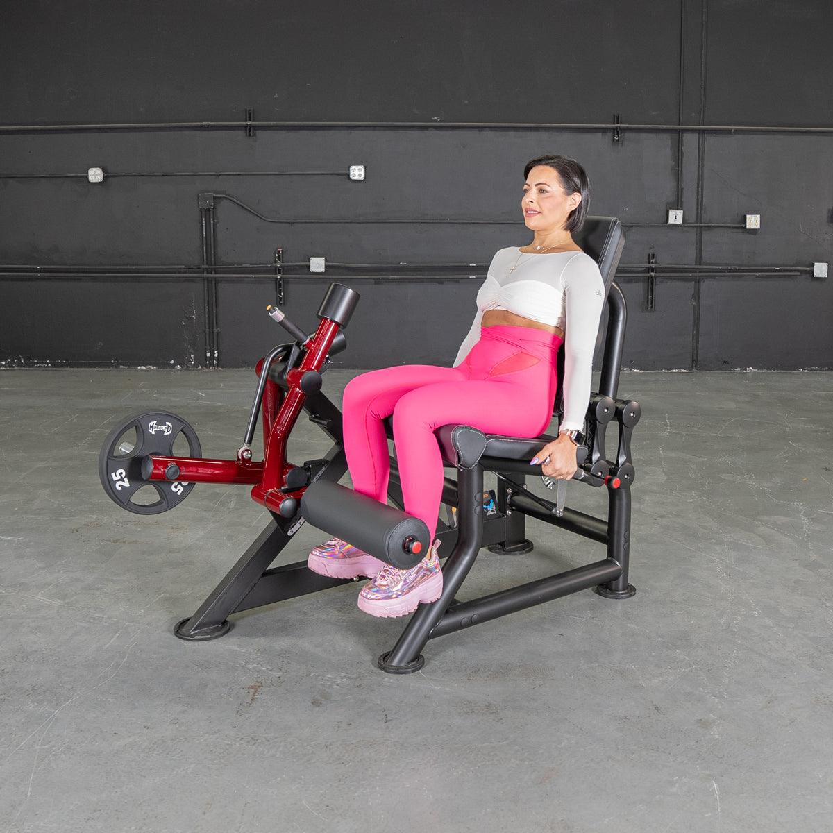 Woman in pink leggings using a red and black leg extension machine with a 25lb weight plate.