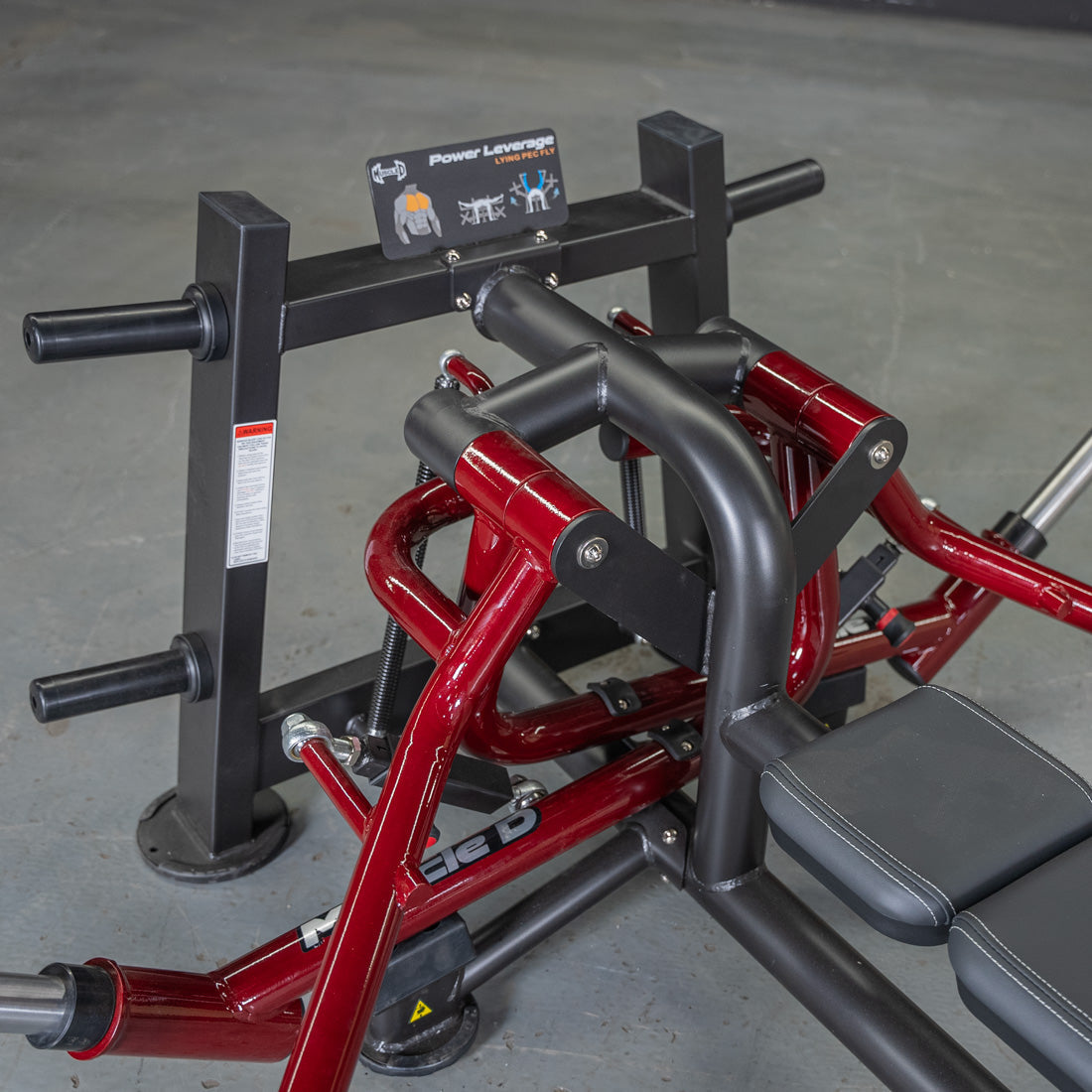 Close-up of a red and black Power Leverage V2 Lying Pec Fly machine with weight plate holders and padded seat.