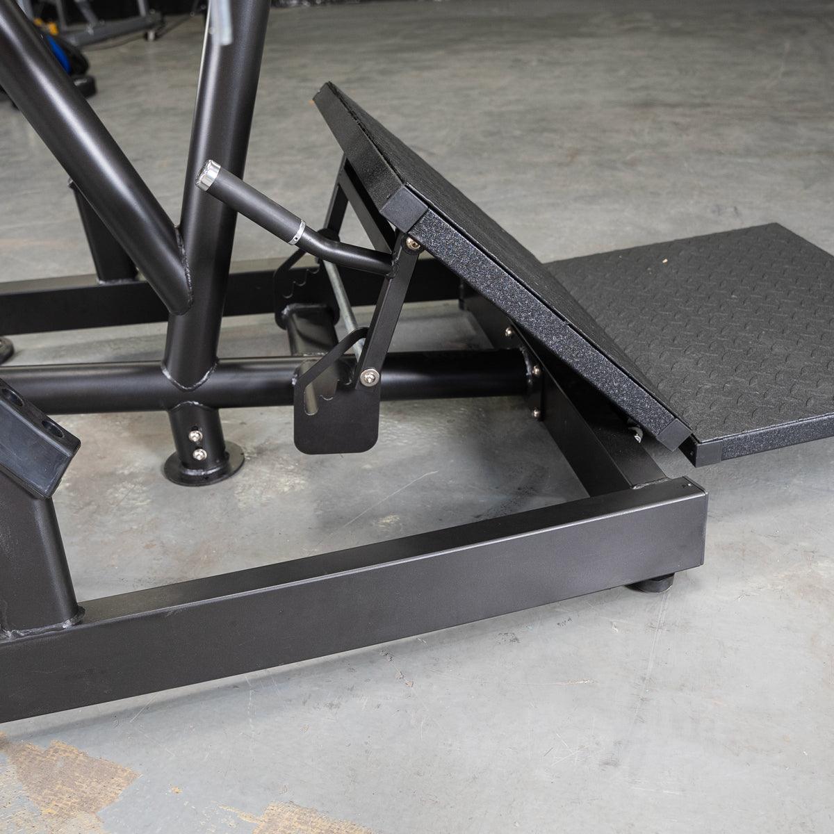 Close-up of the black footplate and base of a pendulum squat machine on a concrete floor.