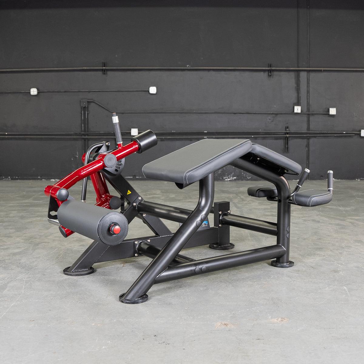 Black and red prone leg curl machine with a padded bench and rollers in a gym setting.