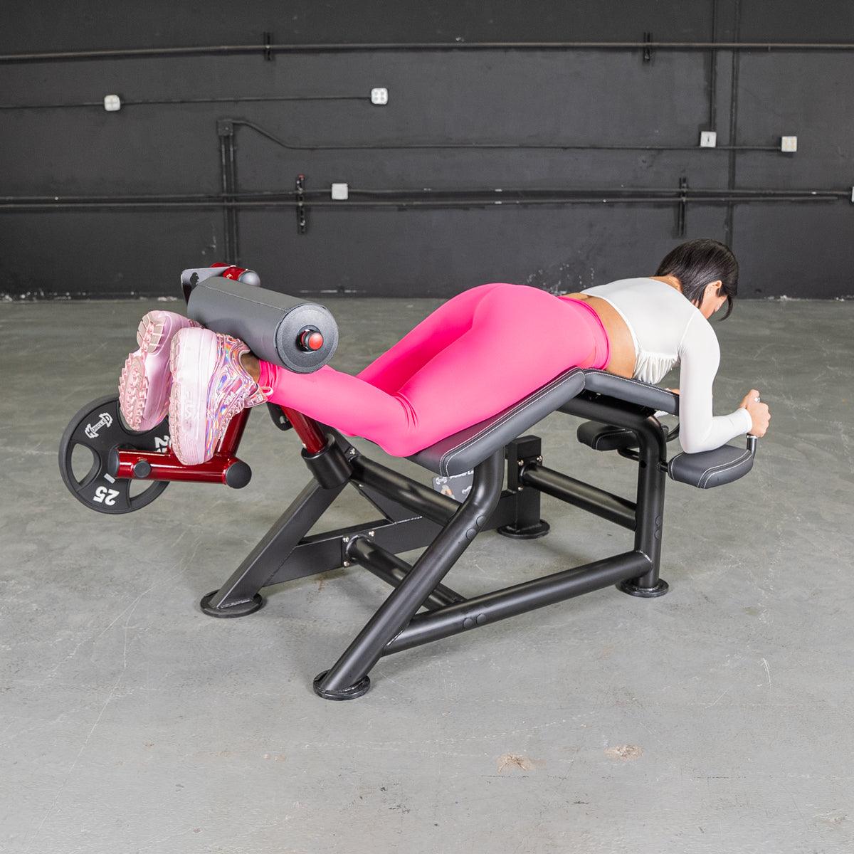 Woman in pink leggings using a prone leg curl machine with a 25lb weight plate.