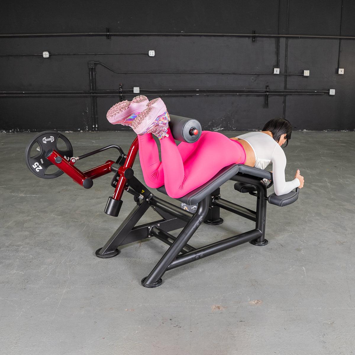 Woman in pink leggings using a red and black prone leg curl machine with a 25lb weight plate.