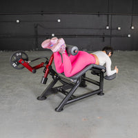 Thumbnail for Woman in pink leggings using a red and black prone leg curl machine with a 25lb weight plate.