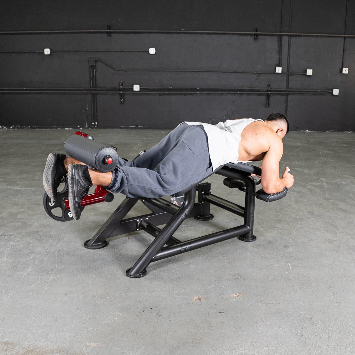 Man using a black and red prone leg curl machine with a weight plate in a gym.