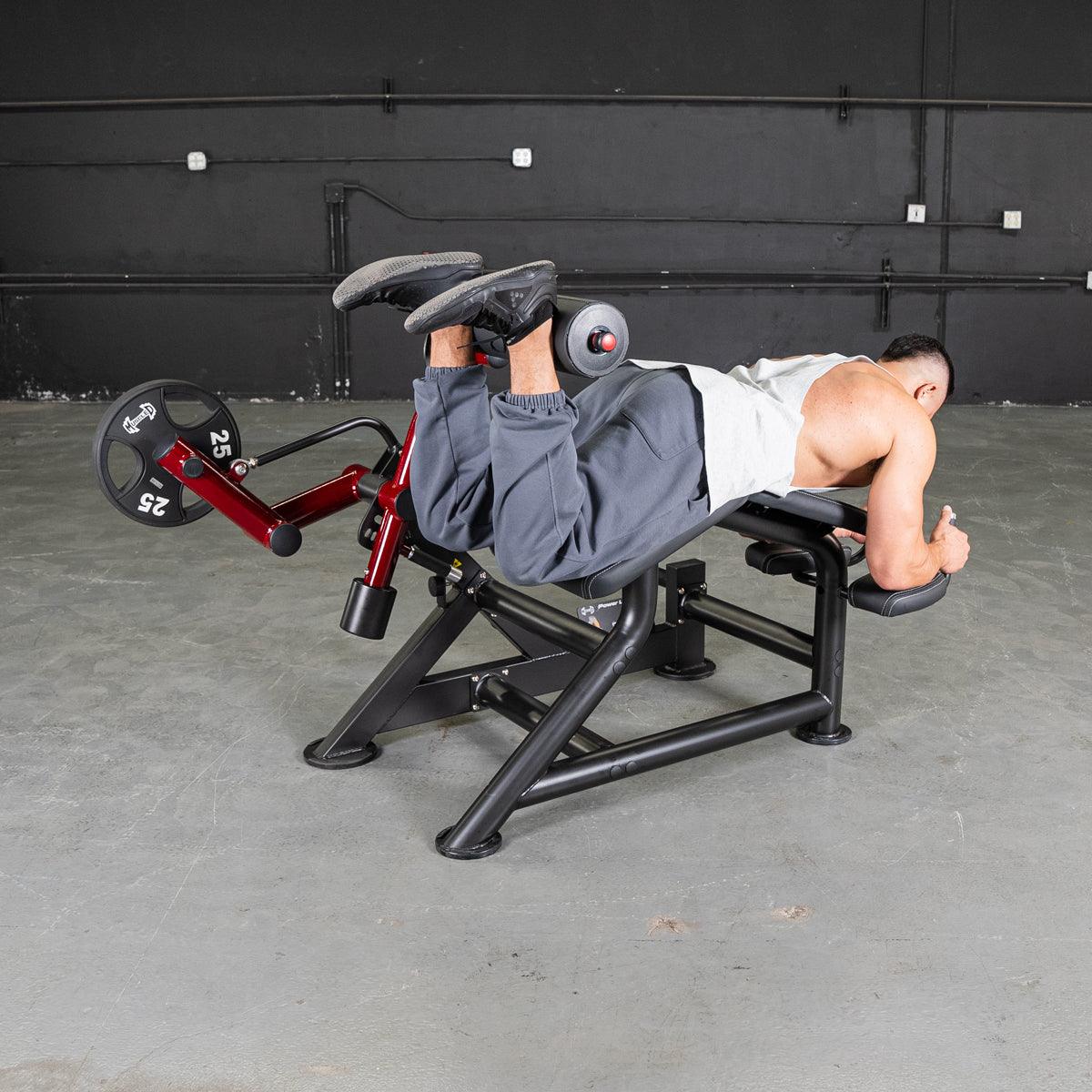 Man performing prone leg curls on a red and black fitness machine with weight plates.