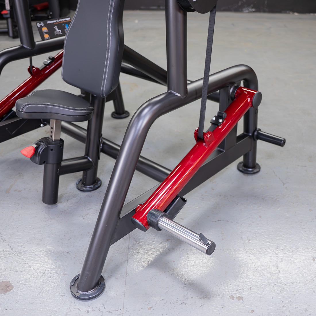 Close-up of a black and red fitness machine with a padded seat and a metal footrest on a concrete floor.