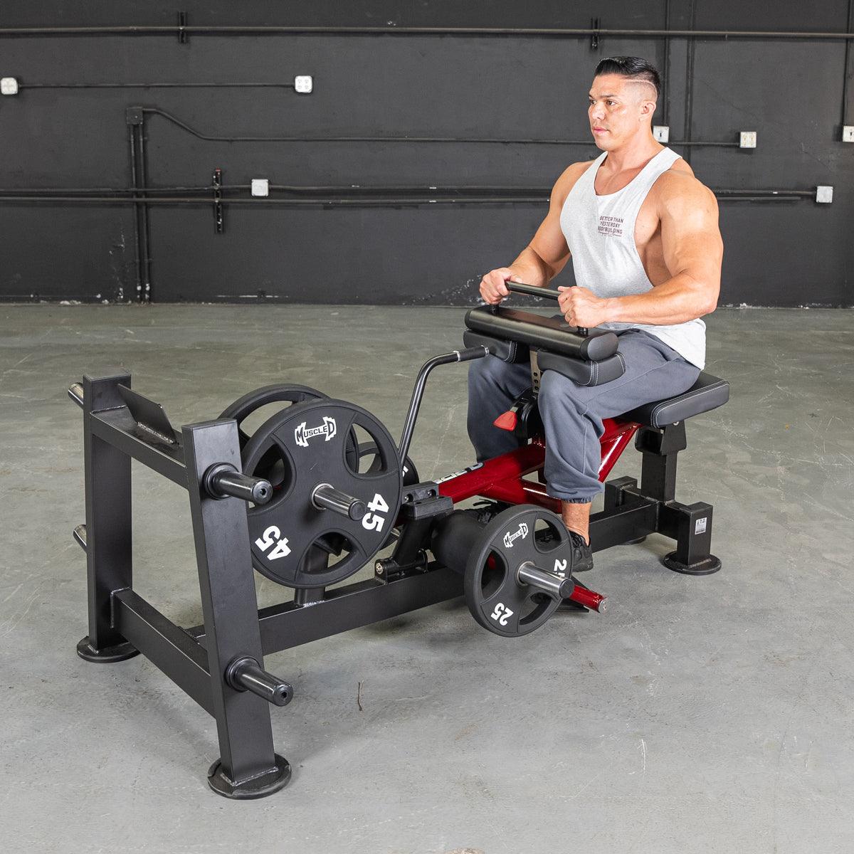 Man seated on a red and black calf/tib machine with weight plates in a gym.