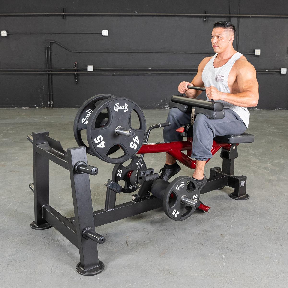 Man in white tank top using a red and black seated calf/tib machine with weights in a gym.