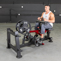 Thumbnail for Man in white tank top using a red and black seated calf/tib machine with weights in a gym.