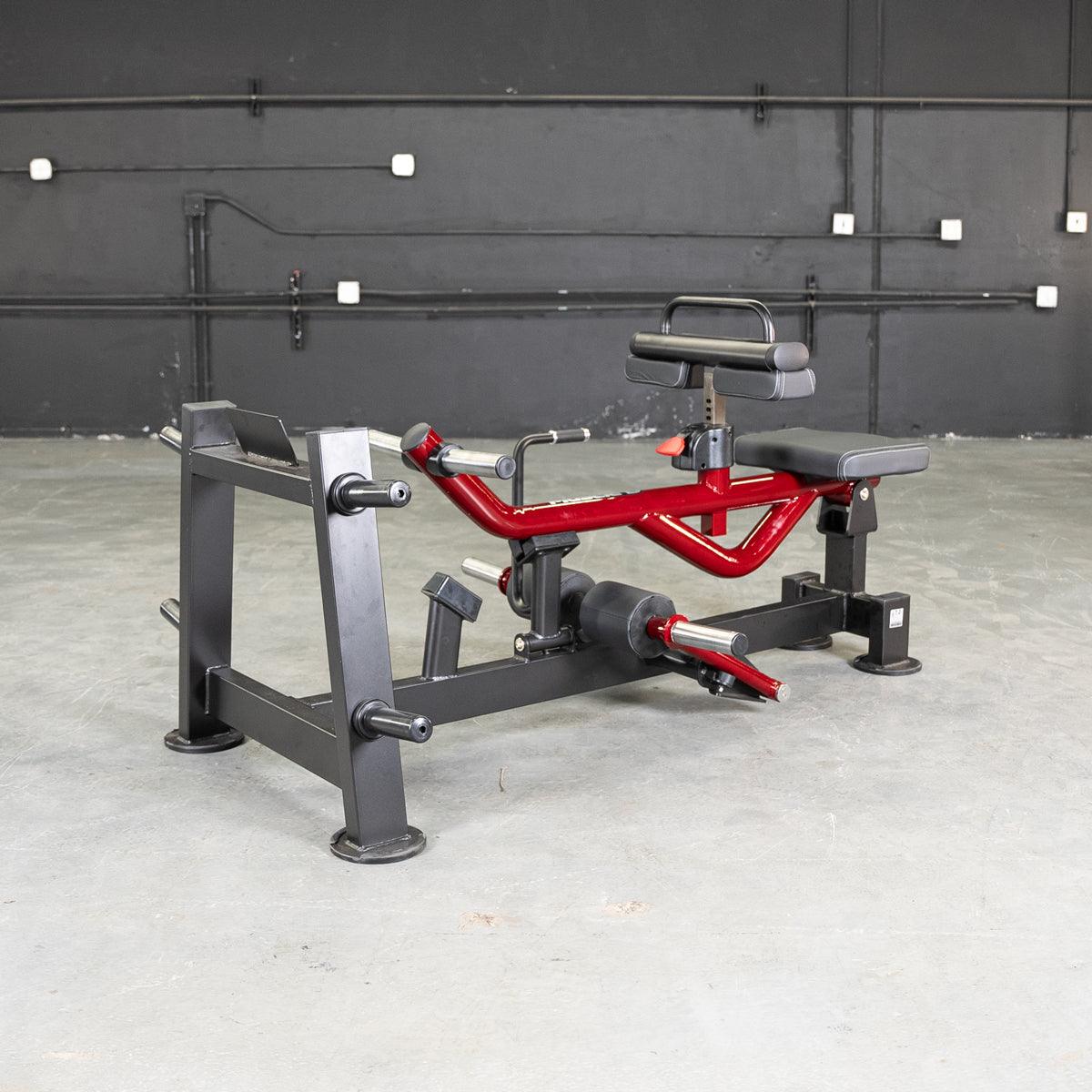 Red and black seated calf/tib machine with padded seat and knee rests, on a concrete floor.