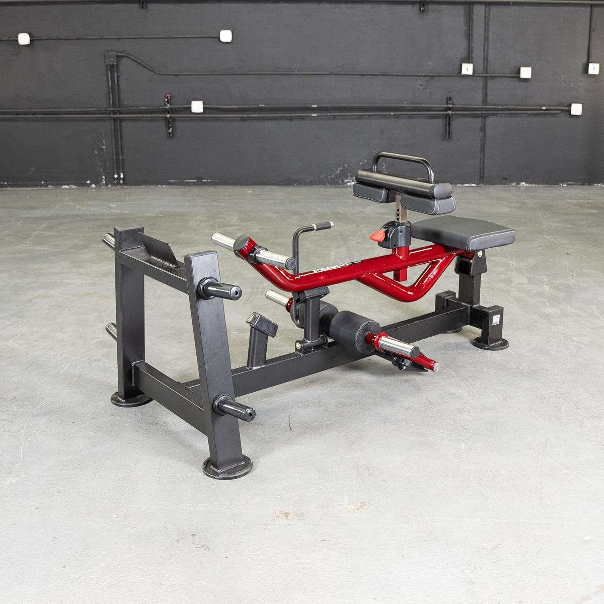 Red and black seated calf/tib machine with padded seat and leg rests on a concrete floor.