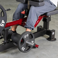 Thumbnail for Person seated on a red and black calf machine with weight plates, performing a calf exercise.