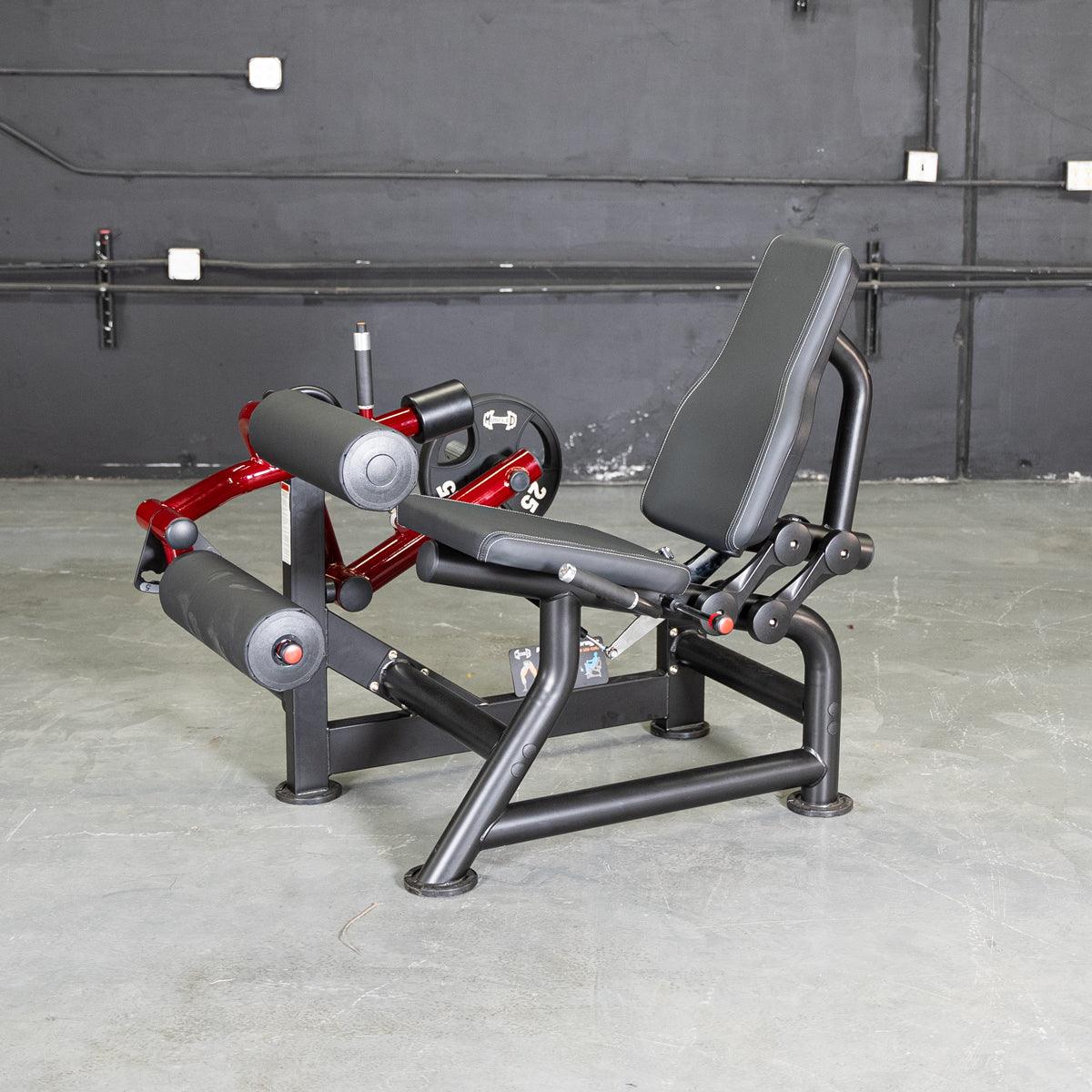 Black and red seated leg curl machine with black padded seat and rollers on a concrete floor.