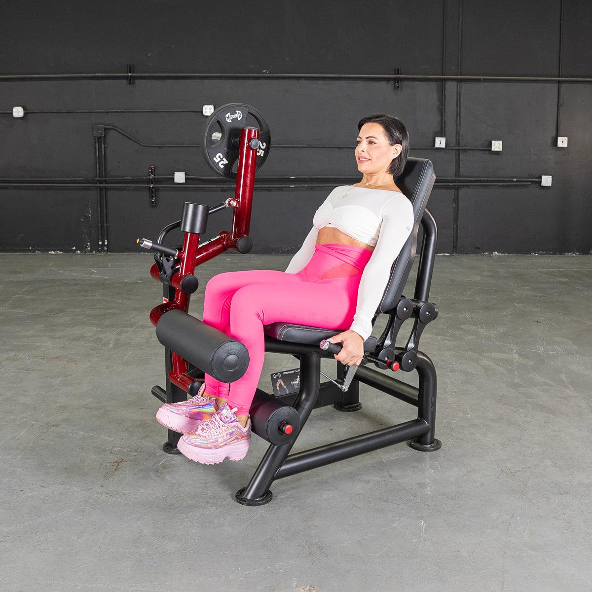 Woman in pink leggings using a red and black seated leg curl machine with weight plates.