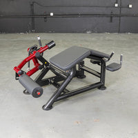 Thumbnail for Red and black prone leg curl machine with padded seat and leg rollers on a concrete floor.