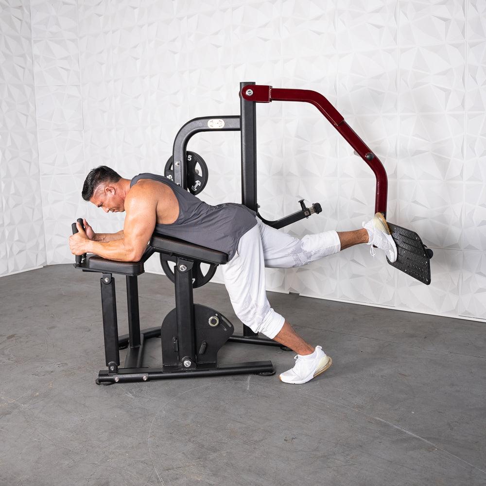 Man using a black and red plate-loaded glute kickback machine, lying prone with one leg extended.