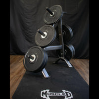 Thumbnail for Black bumper plates on a weight rack with a Muscle D Fitness logo mat.